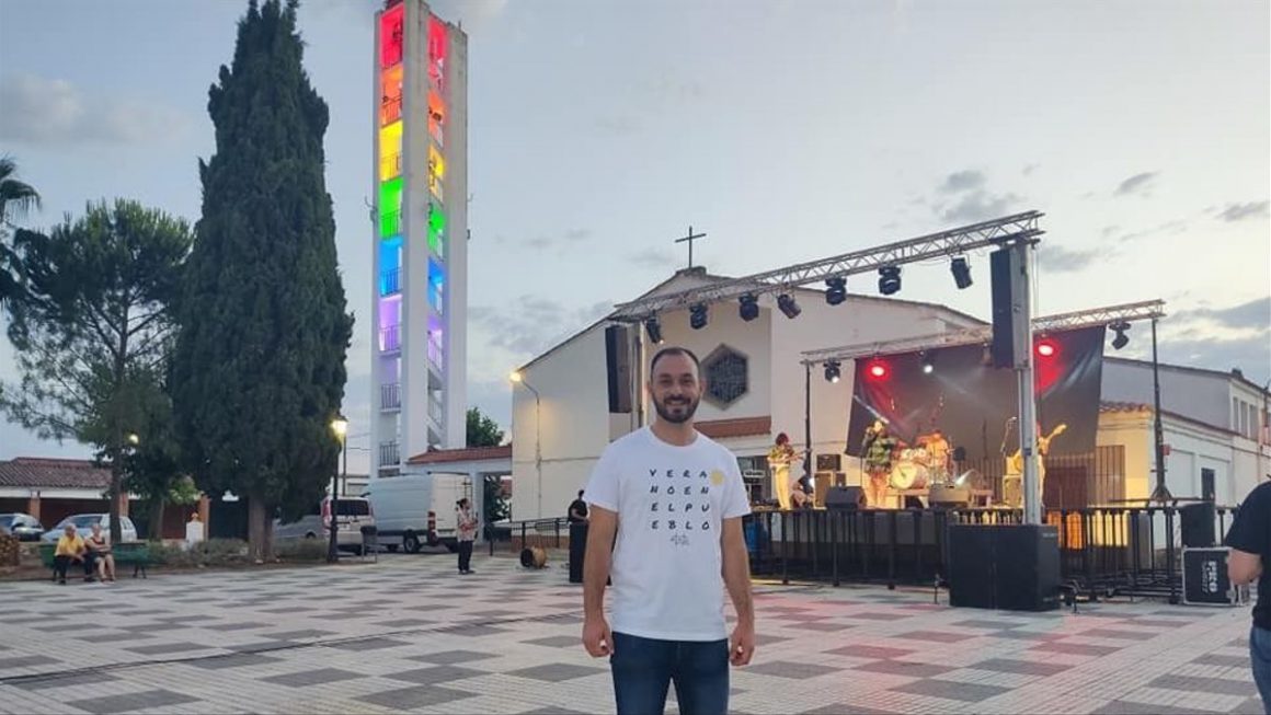 El alcalde de la entidad local de Vivares (Badajoz), Jairo Pino, delante de la iglesia iluminada con los colores de la bandera LGTBI. Imagen Cedida